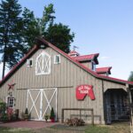 A charming barn at Red Roof Ranch with beige siding and a red roof, featuring a decorative red horse sign. The barn is surrounded by trees, and the entrance is adorned with potted plants and rustic décor.