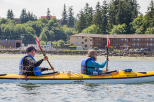 Two kayakers paddle in tandem during Ride the Tide 2024 in Port Orchard, both wearing blue life vests and using red paddles. The yellow kayak glides through the water, with splashes visible, and lush green trees and waterfront buildings are seen in the background.