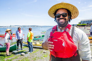 A participant at Ride the Tide 2024 at the Port Orchard waterfront, smiling and wearing a red life vest and straw hat. In the background, other kayakers and paddlers gather near colorful kayaks on a sunny day by the water.