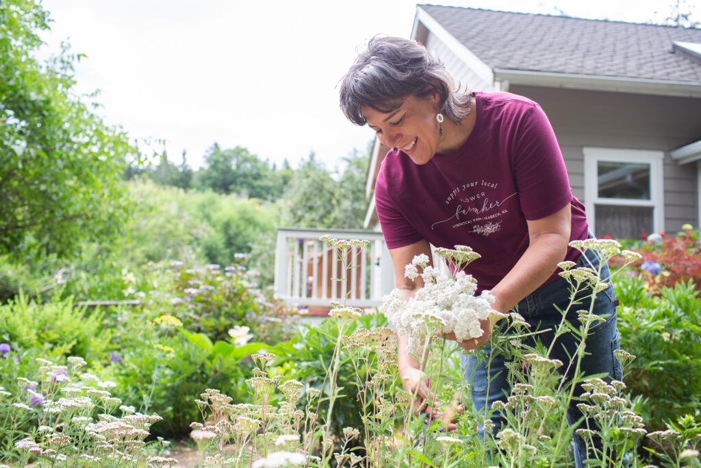 A woman wearing a burgundy T-shirt that reads "Support Your Local Flower Farmer" picks white flowers in a lush garden. The scene features a mix of greenery and colorful blooms, with a house and wooden deck in the background.