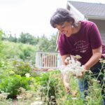 A woman wearing a burgundy T-shirt that reads "Support Your Local Flower Farmer" picks white flowers in a lush garden. The scene features a mix of greenery and colorful blooms, with a house and wooden deck in the background.