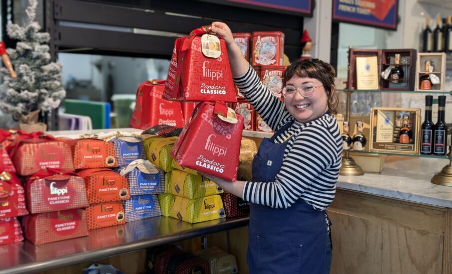 A cheerful woman in glasses and a striped shirt holds two red packages of Filippi Panettone Classico at Via Rosa 11. She is standing behind a festive holiday display featuring colorful boxes of panettone and other Italian holiday treats. A small Christmas tree and decorative items are visible in the background.