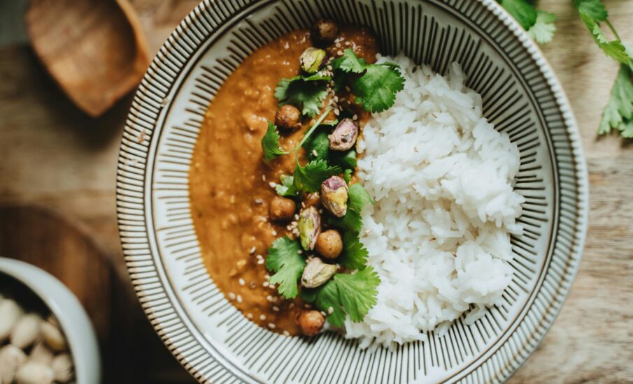 Close-up of a vegan-friendly dish featuring creamy curry topped with roasted chickpeas, pistachios, fresh cilantro, and sesame seeds, served alongside fluffy white rice in a patterned ceramic bowl, perfect for plant-based food lovers