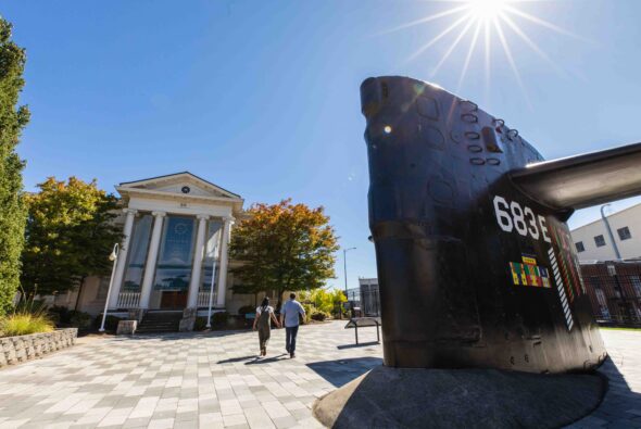 A couple walks hand in hand towards the Puget Sound Navy Museum in Bremerton, WA, with a bright sun overhead. In the foreground, a large black submarine sail with identification markings stands as part of an outdoor exhibit, symbolizing the region's rich naval heritage. Lush trees and a blue sky frame the scene.