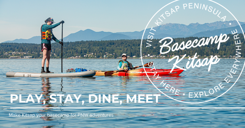 A person paddles a stand-up paddleboard beside another person in a red kayak on calm water with forested hills and the Olympic Mountains in the background. The large white circular logo reads “Basecamp Kitsap – Visit Kitsap Peninsula – Stay Here • Explore Everywhere.” Bold white text says “Play, Stay, Dine, Meet,” with a subheading that reads, “Make Kitsap your basecamp for PNW adventures.”