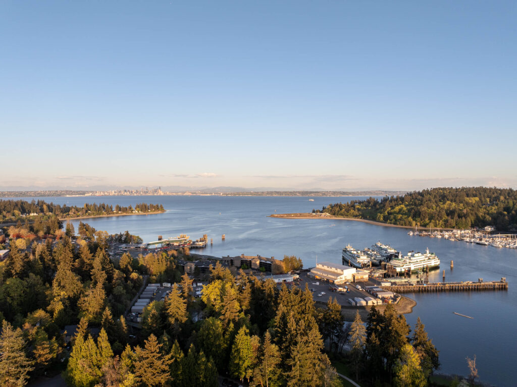 Drone view of downtown Bainbridge Island showcasing its lush greenery, marina filled with ferries and boats, and expansive waterfront, with the skyline of Seattle visible in the distance across Puget Sound under a clear blue sky.