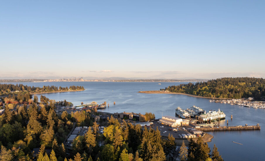 Drone view of downtown Bainbridge Island showcasing its lush greenery, marina filled with ferries and boats, and expansive waterfront, with the skyline of Seattle visible in the distance across Puget Sound under a clear blue sky.