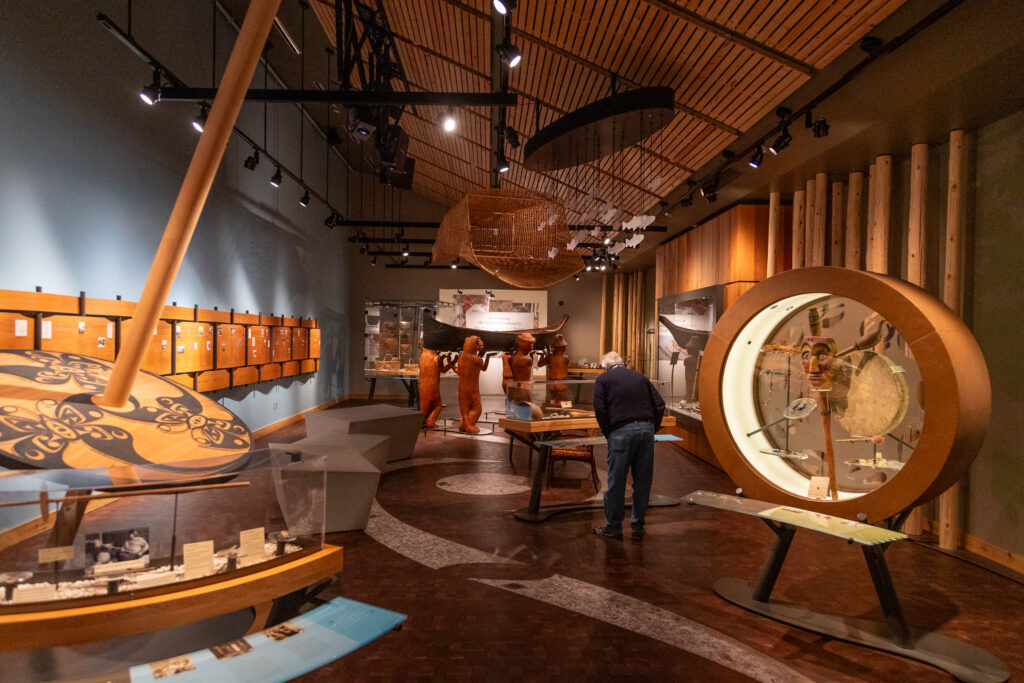 An interior view of a museum showcasing Indigenous art and artifacts, including a carved canoe, traditional masks, intricate displays, and educational panels under warm, modern lighting.