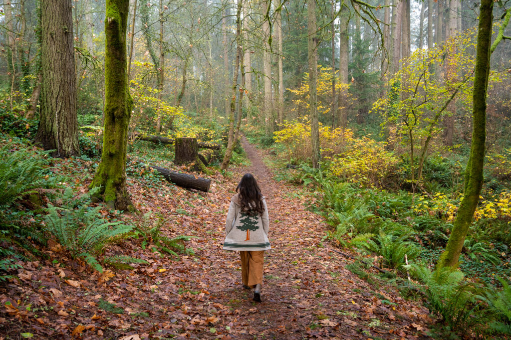 A woman walks along a leaf-covered forest trail surrounded by vibrant autumn foliage, lush green ferns, and moss-covered trees, embracing the peaceful woodland atmosphere.