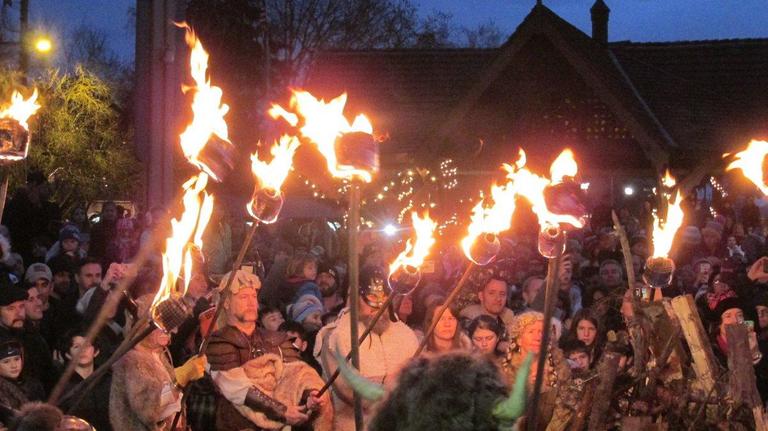 A vibrant crowd gathers at dusk in Poulsbo, WA, for a Viking-themed bonfire celebration. Participants dressed in Nordic-inspired attire hold flaming torches high, illuminating the festive atmosphere. The event, rich in tradition, reflects Poulsbo’s Scandinavian heritage and community spirit.