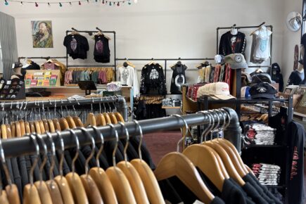 Interior of Fingers Duke, a local art and apparel shop in Bremerton, WA. The store features racks of graphic t-shirts, hoodies, hats, and accessories, with colorful string lights overhead. Art prints, cards, and custom designs line the shelves, creating a vibrant and creative atmosphere. A portrait painting and screen-printed apparel add to the artistic vibe of the space.