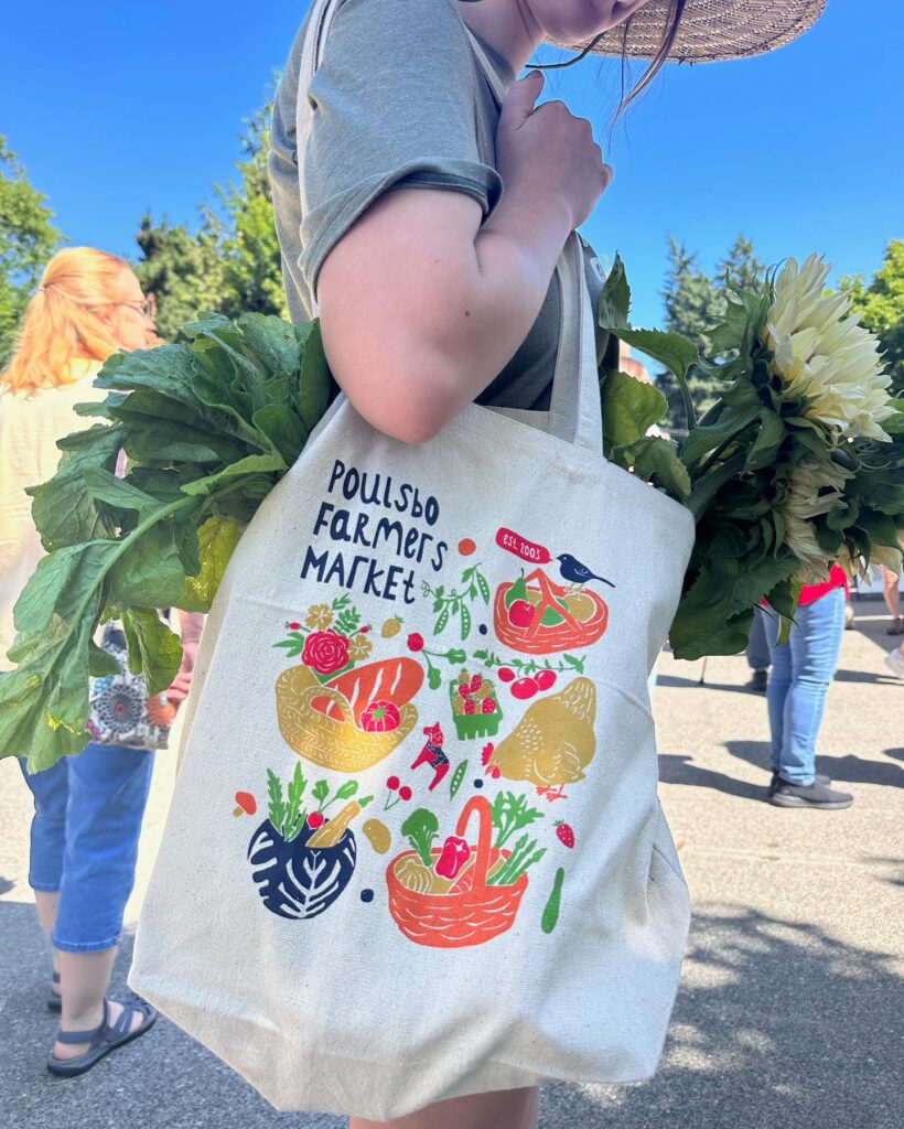 A person wearing a wide-brimmed straw hat holds a canvas tote bag filled with leafy greens and white flowers at an outdoor market. The tote bag features colorful illustrations of baskets filled with produce, a chicken, flowers, and a bird, with the text "Poulsbo Farmers Market" and "EST. 2003" printed on it. Other shoppers and a clear blue sky are visible in the background.