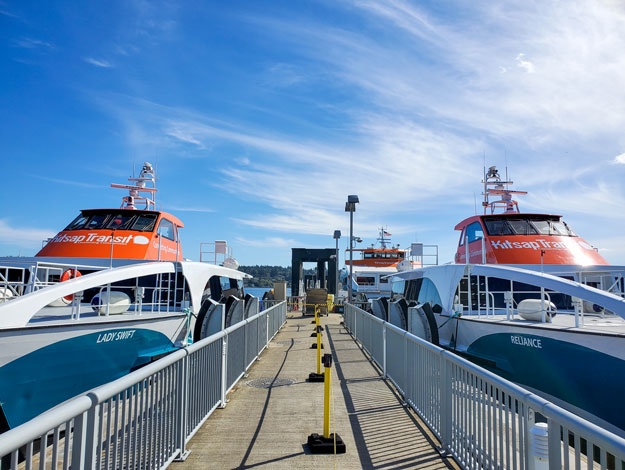 Kitsap Transit fast ferries, Lady Swift and Reliance, docked side by side at a terminal under a bright blue sky, ready for passenger boarding.