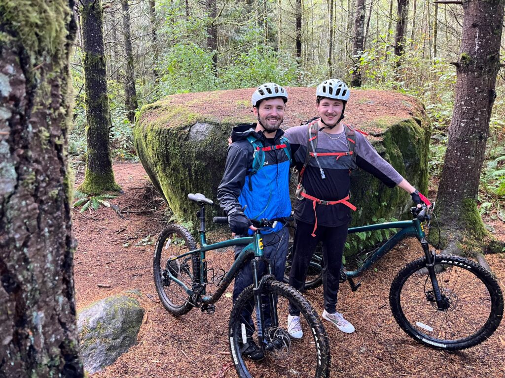 Two smiling cyclists wearing helmets and backpacks pose with muddy mountain bikes in a mossy forest next to a massive, lichen-covered boulder on a Kitsap Peninsula trail.