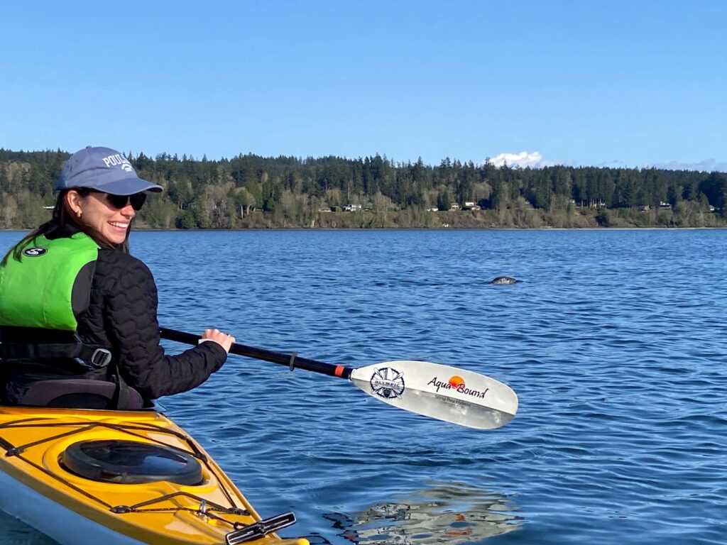 A woman in a kayak paddles on calm blue water under a clear sky, smiling as a harbor seal surfaces in the distance near Poulsbo.