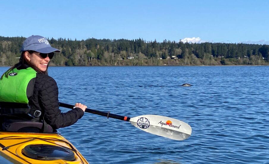 A woman in a kayak paddles on calm blue water under a clear sky, smiling as a harbor seal surfaces in the distance near Poulsbo.