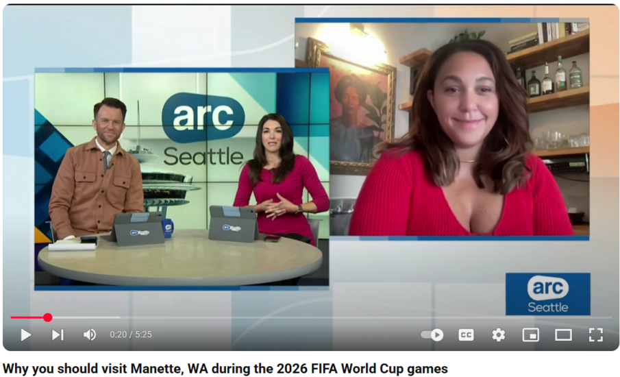 Screenshot of a KOMO4 ARC Seattle interview. On the left, two news anchors sit at a studio desk in front of a large digital screen displaying the "arc Seattle" logo. On the right, Anna Bressi, owner of Lola’s Cafe in Manette, WA, appears on a split-screen video call. She is smiling and seated in front of a wall with floating shelves, bottles, and a framed portrait behind her. The lower third caption reads: "Why you should visit Manette, WA during the 2026 FIFA World Cup games.