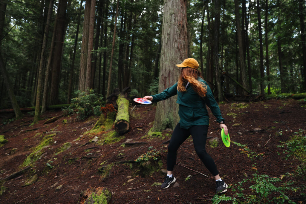 A woman in outdoor gear throws a disc in a wooded disc golf course surrounded by tall trees and mossy logs.