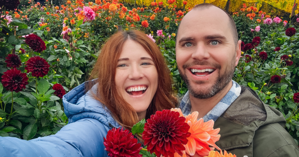 A smiling couple stands in a vibrant dahlia field, holding colorful blooms. The woman wears a blue puffy jacket and the man a green coat. Rows of red, orange, and pink flowers stretch behind them under an overcast sky with evergreens in the distance.