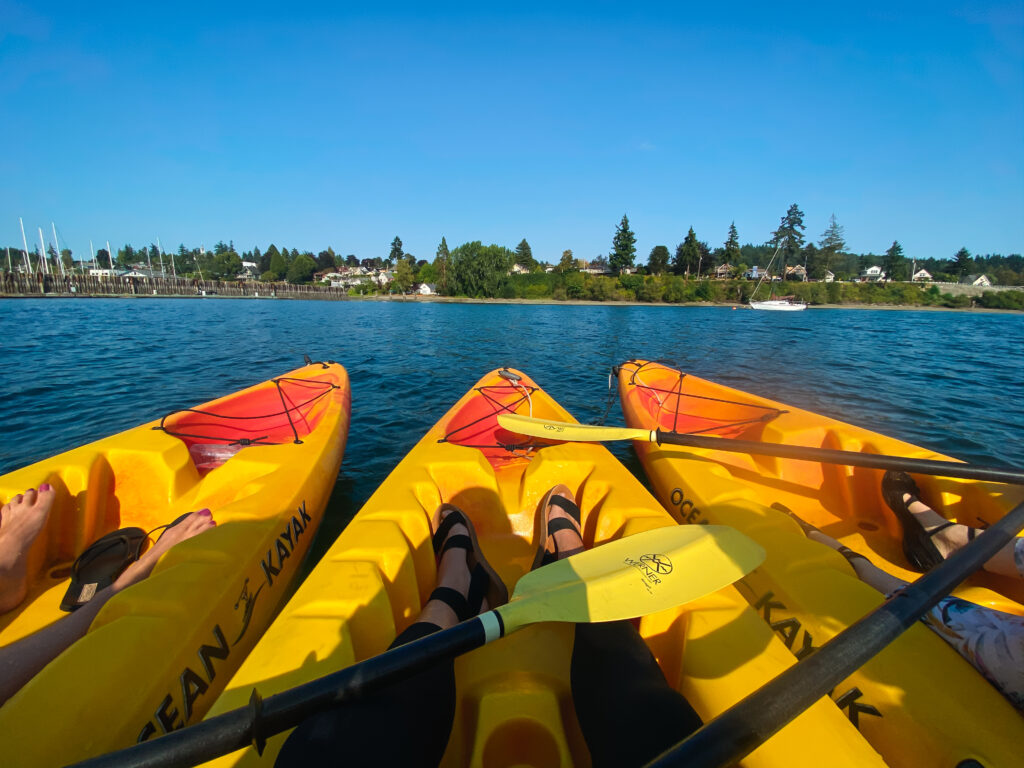 Three bright yellow ocean kayaks with paddlers gliding on blue water, facing the tree-lined shoreline and docked sailboats under a clear summer sky.