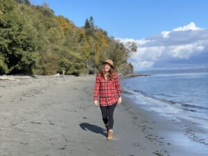 A woman in a red flannel shirt and hat walks along a sandy shoreline with a forested backdrop and cloudy skies.