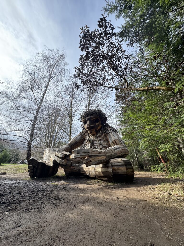 A large whimsical wooden troll sculpture named Pia the Peacekeeper, seated in a wooded clearing surrounded by trees and muddy forest paths.