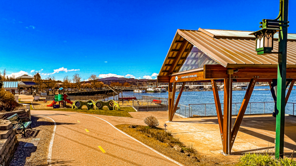 A vibrant waterfront park at the Port Orchard Marina featuring a covered overlook structure, children's playground, and walking path with views of boats docked on the water under a bright blue sky.
