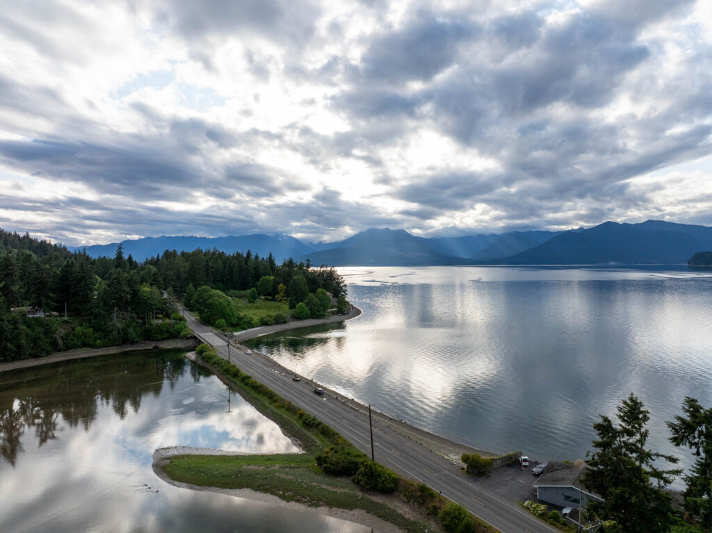  dramatic aerial view of a coastal road in Kitsap Peninsula, lined by lush forest on one side and calm reflective waters on the other, with the Olympic Mountains silhouetted in the distance under a cloudy sky.