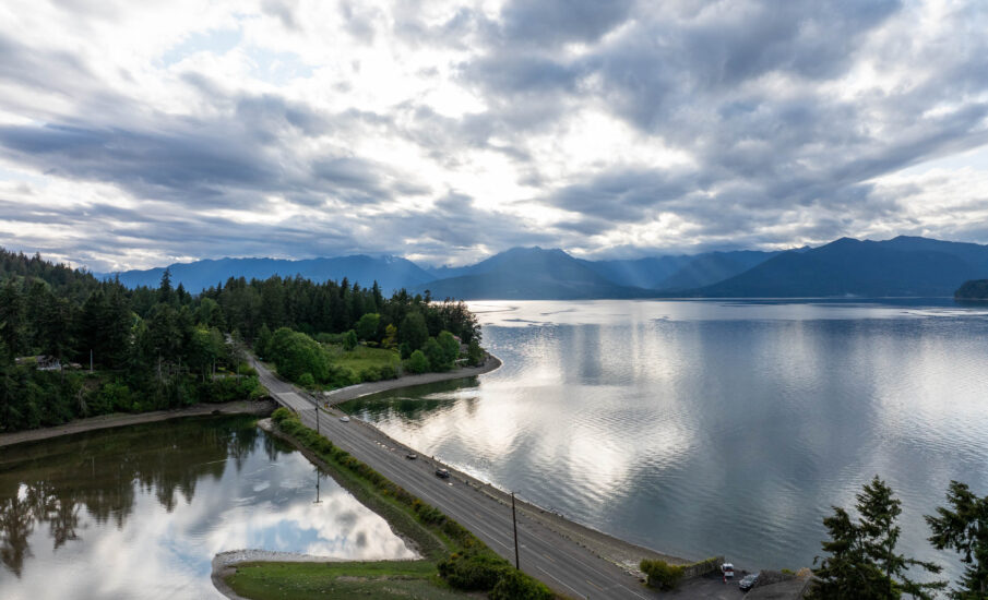 dramatic aerial view of a coastal road in Kitsap Peninsula, lined by lush forest on one side and calm reflective waters on the other, with the Olympic Mountains silhouetted in the distance under a cloudy sky.