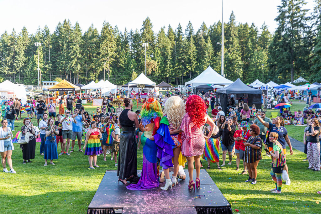 A vibrant crowd gathers on the lawn at Kitsap Pride, watching drag performers on stage under sunny skies and surrounded by vendor tents and forest trees.