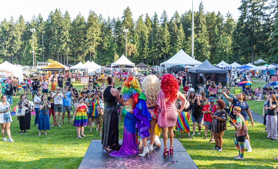 A vibrant crowd gathers on the lawn at Kitsap Pride, watching drag performers on stage under sunny skies and surrounded by vendor tents and forest trees.