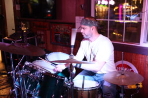 Drummer performing onstage at a local Bremerton bar, immersed in live music under warm lighting with music equipment in the background.