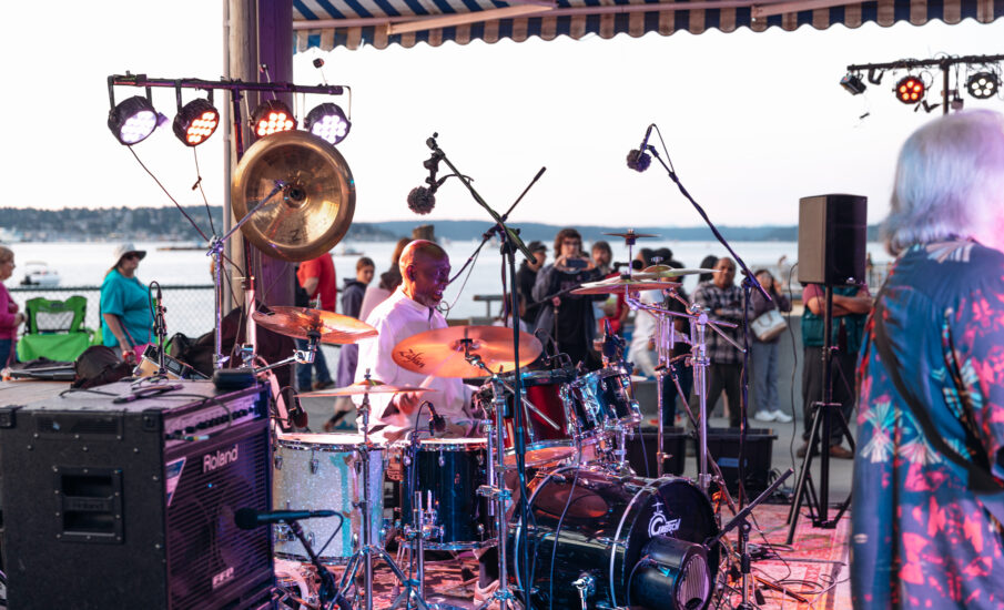 A Black male drummer energetically performs on a waterfront stage at sunset, surrounded by drum equipment, lighting rigs, and an engaged audience in the background.
