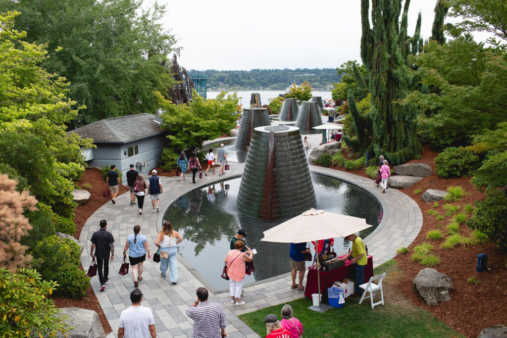 Visitors stroll through the Bremerton Harborside Fountain Park during the Kitsap Wine Festival, surrounded by modern fountains, lush greenery, and vendor tents with a view of the Puget Sound.