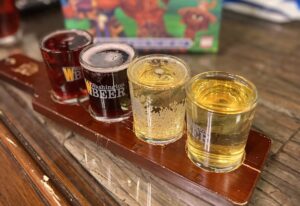 Craft beer flight with four samples on a wooden tray in front of a colorful board game box titled “Fantahzee: Hordes & Heroes,” set at Ashley’s Pub.