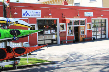 The exterior of Olympic Outdoor Center in Port Gamble, WA, features a bright red historic firehouse building with large garage-style windows and signage above the doors. A kayak trailer with colorful kayaks is parked out front, ready for adventure. The storefront sits on a quiet street lined with charming architecture.