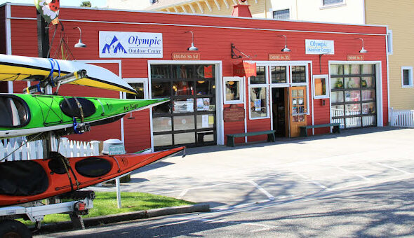 The exterior of Olympic Outdoor Center in Port Gamble, WA, features a bright red historic firehouse building with large garage-style windows and signage above the doors. A kayak trailer with colorful kayaks is parked out front, ready for adventure. The storefront sits on a quiet street lined with charming architecture.