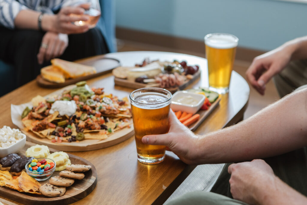 A close-up of hands enjoying pints of beer and an assortment of snacks, including nachos, charcuterie, cookies, and veggies at The SIP’s lounge space.