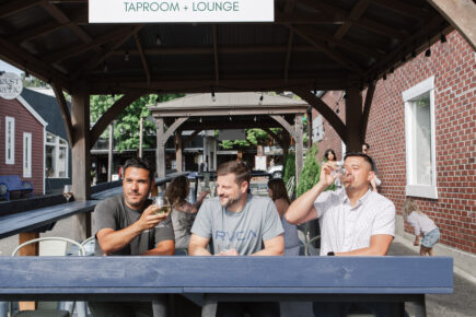 Three men enjoy drinks on the outdoor patio of The SIP Taproom + Lounge in Poulsbo, WA, under a wooden pergola with a sign reading “Pour Your Own.”