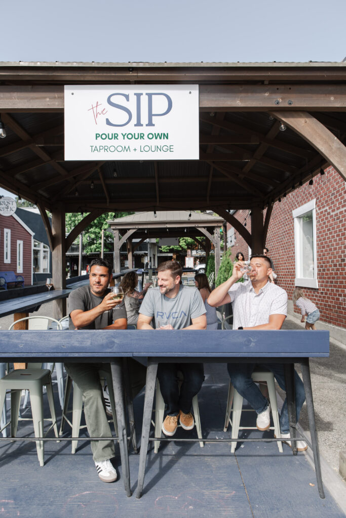 Three men enjoy drinks on the outdoor patio of The SIP Taproom + Lounge in Poulsbo, WA, under a wooden pergola with a sign reading “Pour Your Own.”