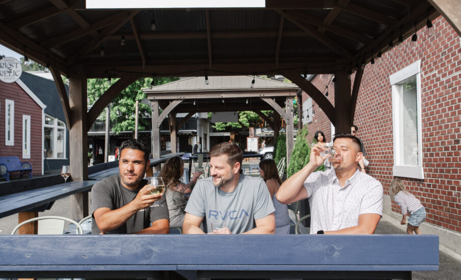 Three men enjoy drinks on the outdoor patio of The SIP Taproom + Lounge in Poulsbo, WA, under a wooden pergola with a sign reading “Pour Your Own.”