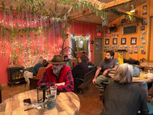 People relax and enjoy drinks inside Bushel & Barrel Ciderhouse in Poulsbo, WA. The cozy interior features warm lighting, hanging greenery, string lights, and rustic wooden tables, creating a welcoming and festive atmosphere. A wall of framed photos and vibrant orange-red tones adds to the ciderhouse’s quirky charm.