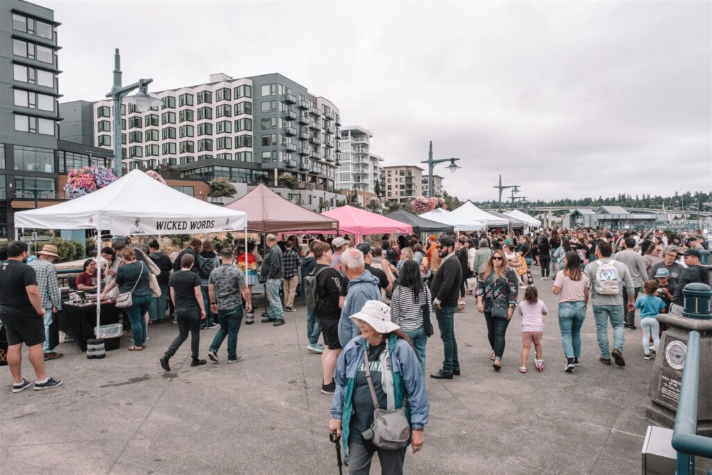 A vibrant street fair scene during the Puget Sound Book Fest in downtown Bremerton, with local vendors, shoppers, and tall modern buildings in the background.