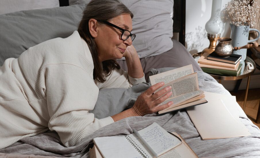 An older woman wearing glasses and a cozy cream-colored robe lies comfortably in bed reading a book. She appears content and relaxed, surrounded by soft pillows, a notebook with handwritten notes, and a stack of books and a vintage lamp on the bedside table.