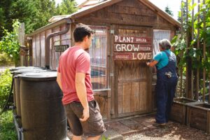 A man and a woman stand outside a rustic wooden greenhouse at The Wanderer’s Nest Therapeutic Farm on Bainbridge Island, WA. The woman, wearing overalls, opens the door next to a sign that reads, “Plant Smiles, Grow Giggles, Harvest Love.” The setting is lush with greenery, garden beds, and rain barrels.Ask ChatGPT
