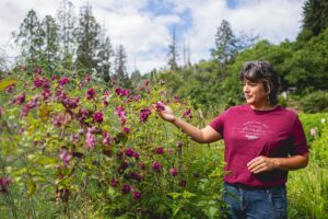 A woman wearing a maroon T-shirt that reads “Support Your Local Flower Farmer – Vinnocki Farm, Seabeck, WA” gently holds a cluster of deep pink blooms in a lush garden. Surrounded by vibrant flowers and greenery, she stands against a backdrop of tall trees and a bright, partly cloudy sky.
