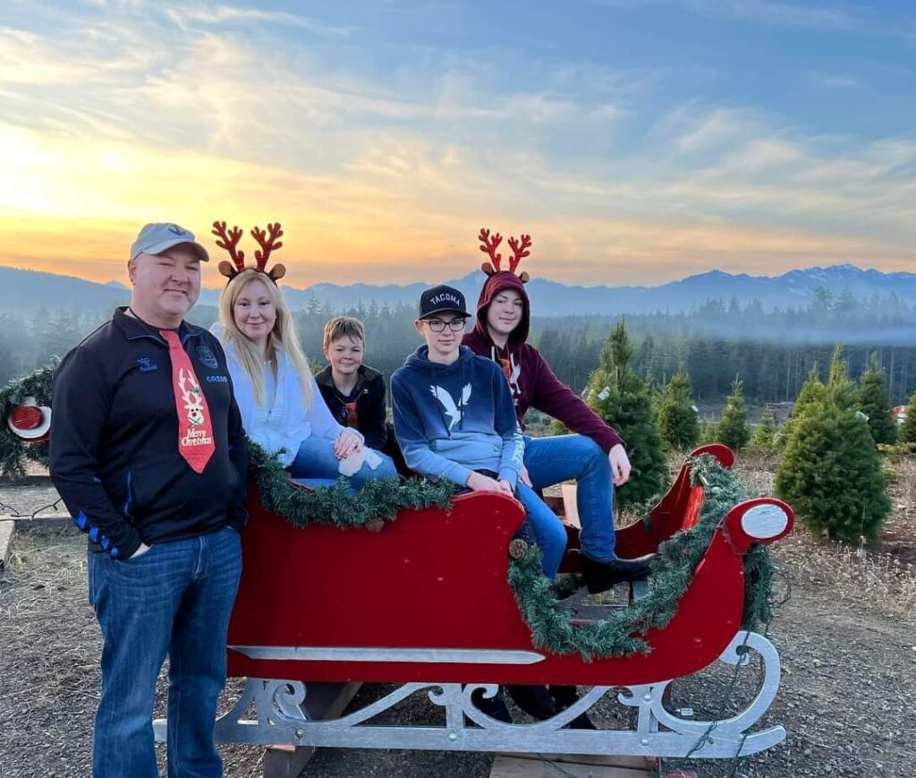 A family poses in a red holiday sleigh at Hubert’s Christmas Tree Farm in Seabeck, framed by evergreens and the Olympic Mountains at sunset.