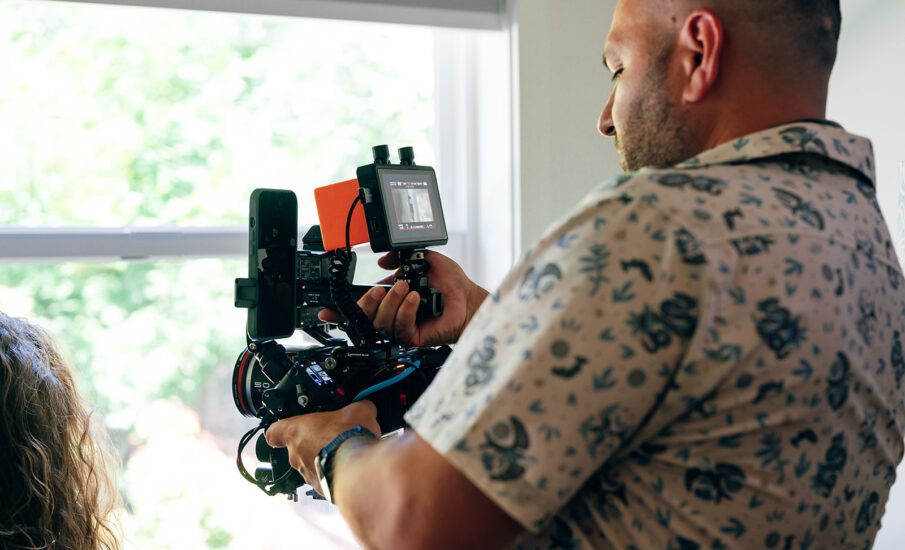 Image shows a man operating a professional video camera indoors near a bright window. He is focused on filming, wearing a light patterned shirt with blue floral designs. The camera is equipped with multiple attachments and a small monitor displaying the shot. Sunlight filters through the window, softly illuminating the scene, suggesting a creative work environment.