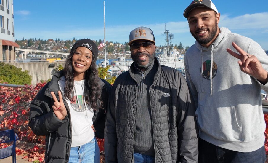 Three people from the Traveling While Black team stand smiling at the Bremerton waterfront on a sunny day. The two on the left and right are holding up peace signs. Fall-colored vines, boats, and part of downtown Bremerton appear in the background.