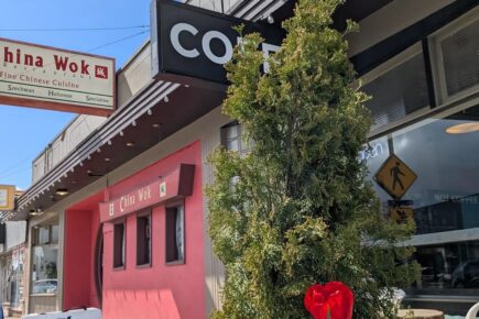 Exterior of Café Corvo on a sunny day, with a hanging café sign, a sidewalk sign reading “Coffee Espresso Pastries,” bright red tulips in the foreground, and neighboring storefronts along a downtown street.